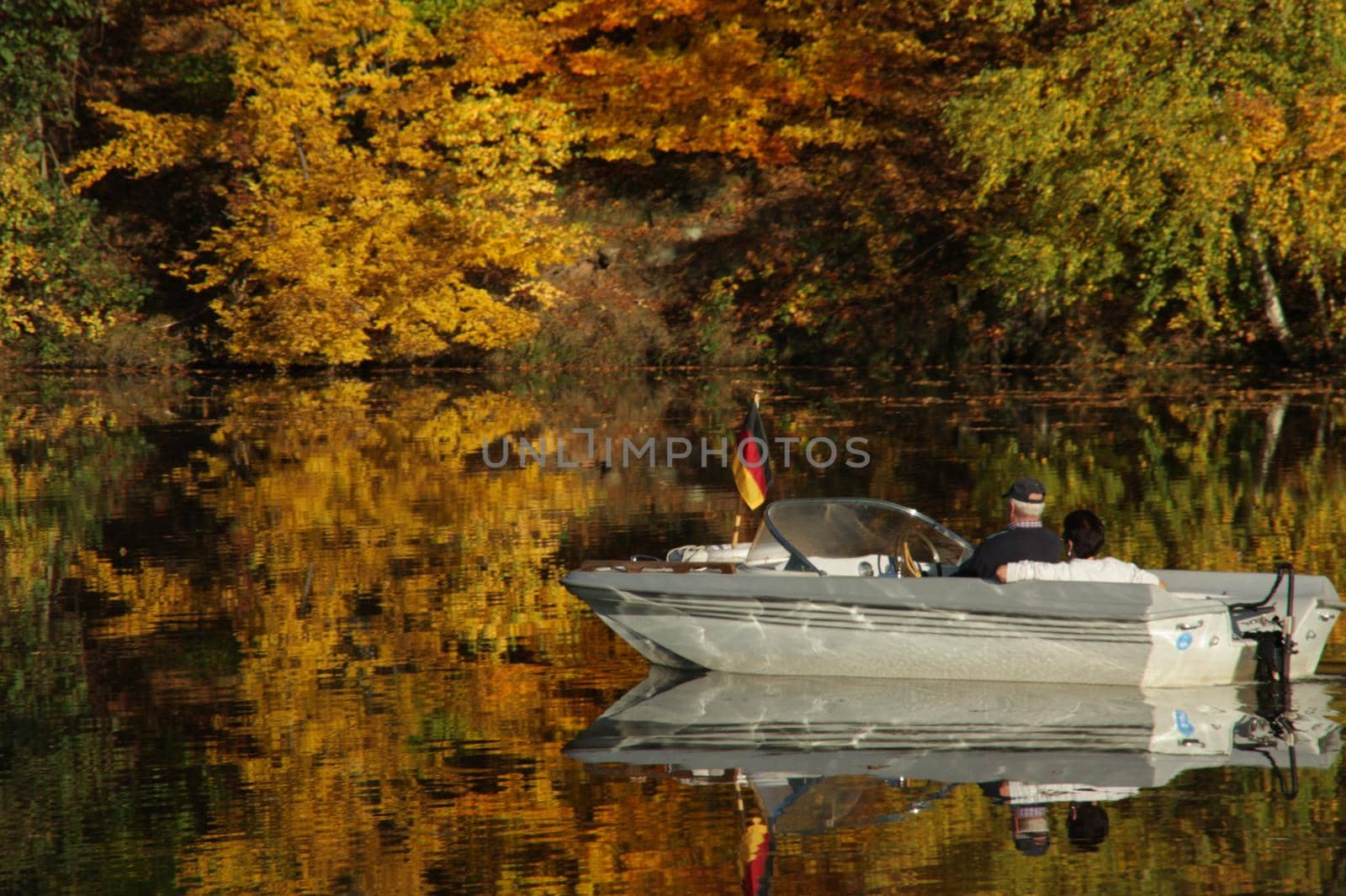 a motorboat and an autumn forest of red yellow and green autumn colors are reflected in the lake surface by Costin