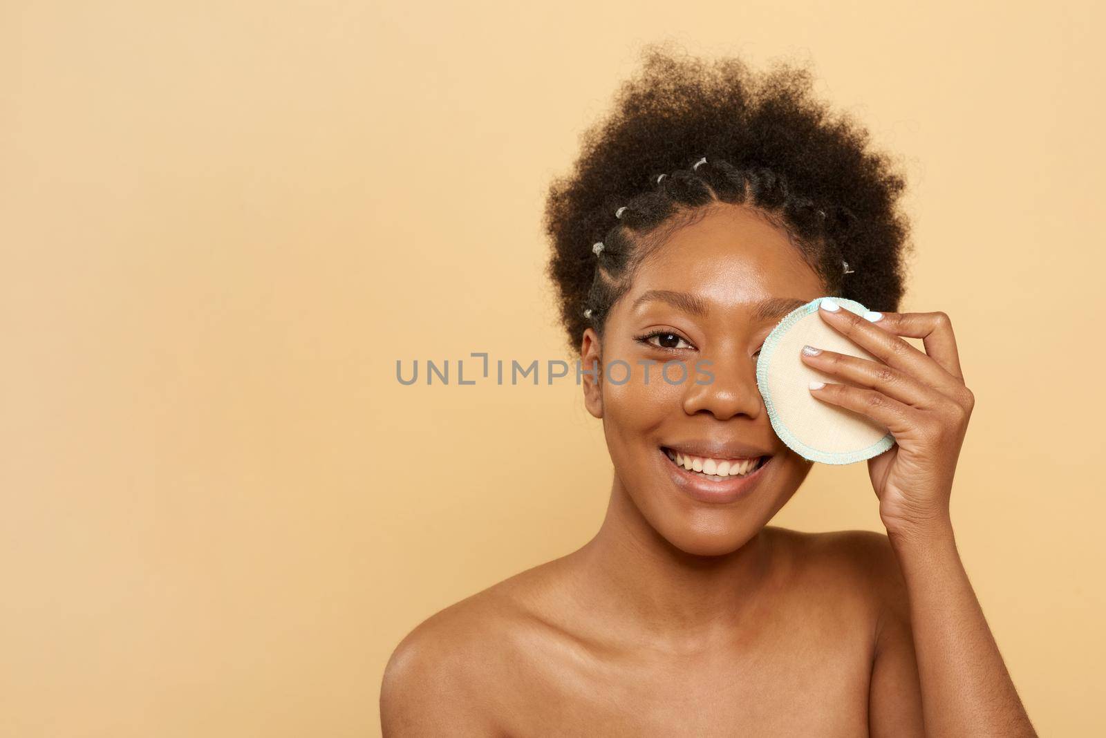 Afro american black young woman holding a reusable skin care cotton pad near her eye, smiling and looking at the camera. Zero waste concept
