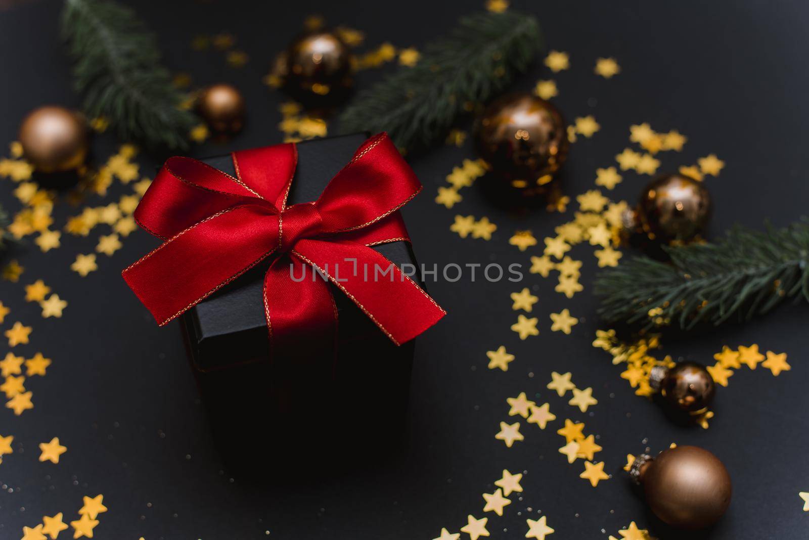 Stylish black gift with a red bow on the background of Christmas decorations, confetti and fir branches by etonastenka