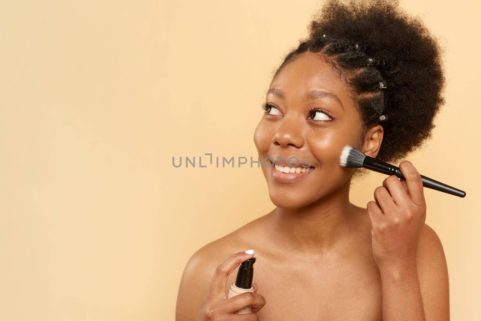 Beautiful smiling woman applying foundation on her face on a beige background. Makeup concept and copy space.