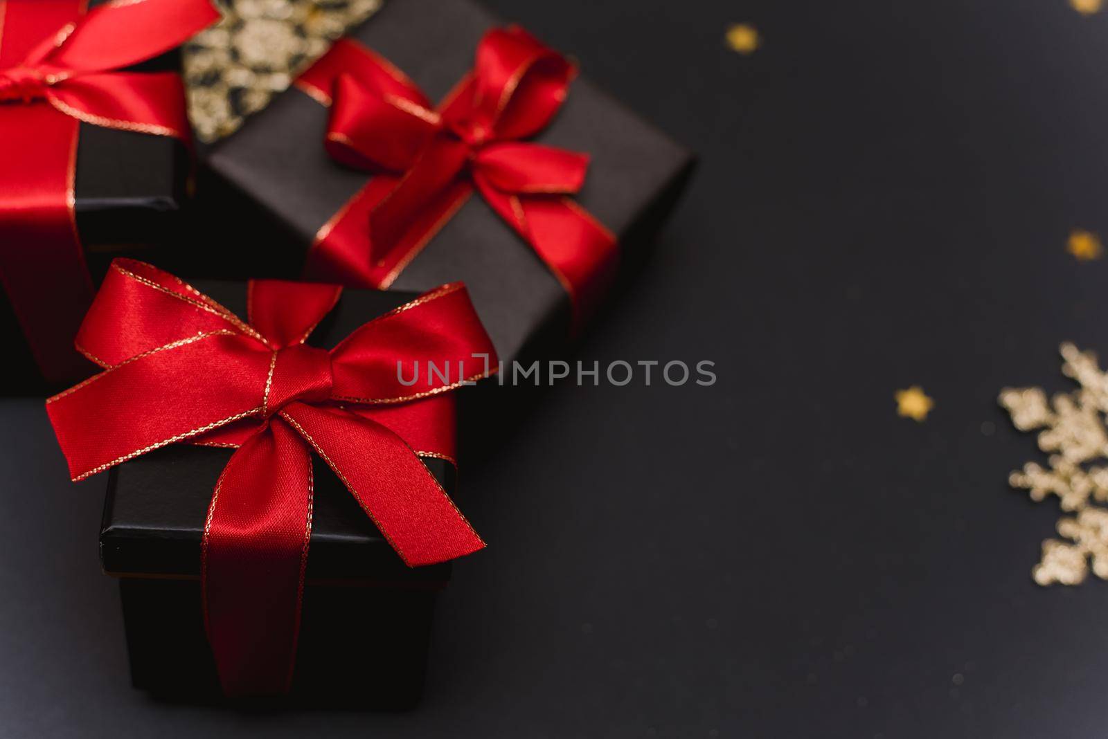 Black gift boxes with a red ribbon on a gold confetti background. Elegant Christmas background with modern golden Christmas decorations, balls, gift boxes, confetti stars on dark black background. Flat design, top view, copy space