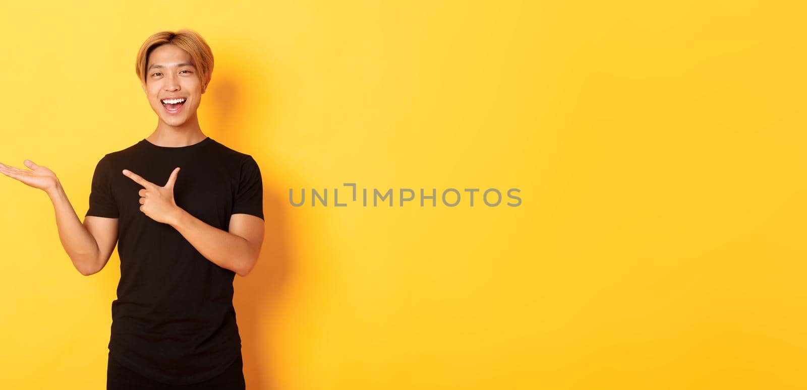 Portrait of handsome smiling asian guy holding something on hand and pointing finger at it, standing yellow background pleased by Benzoix