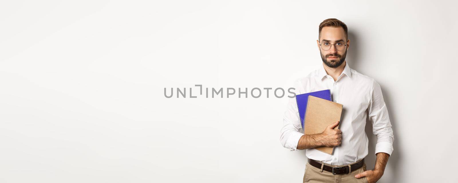 Handsome male employee holding notebooks, looking confident, standing over white background by Benzoix