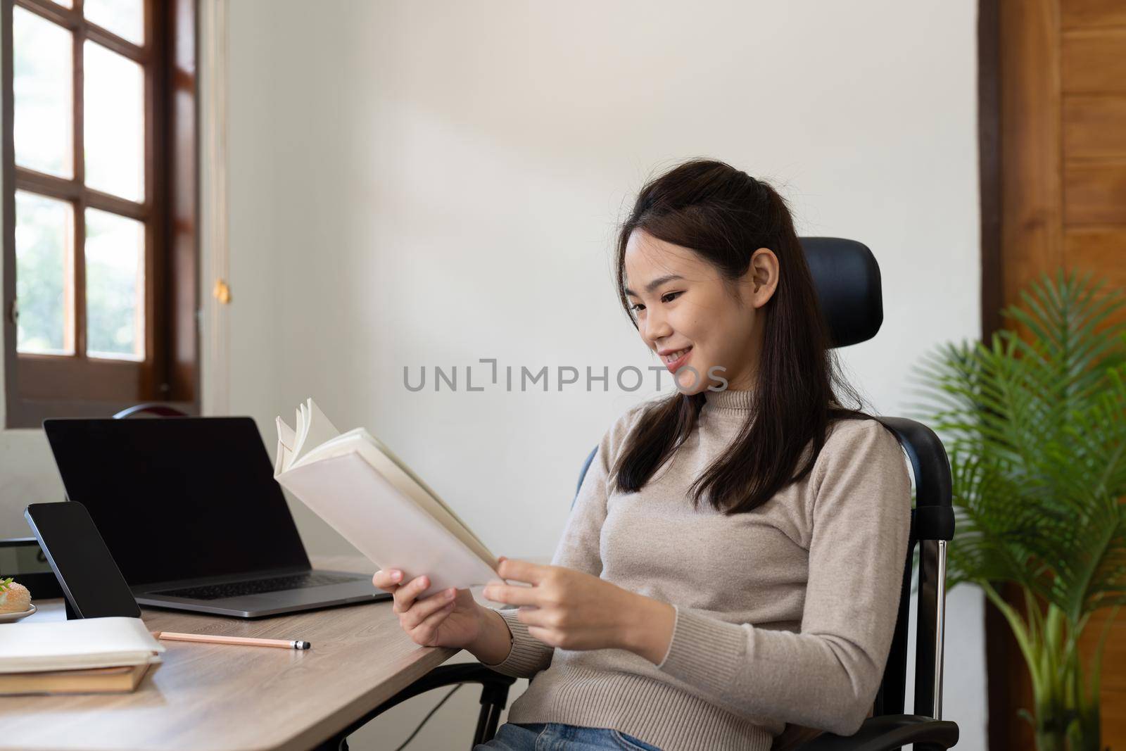 Portrait of asian woman reading a book while studying online learning on laptop at home