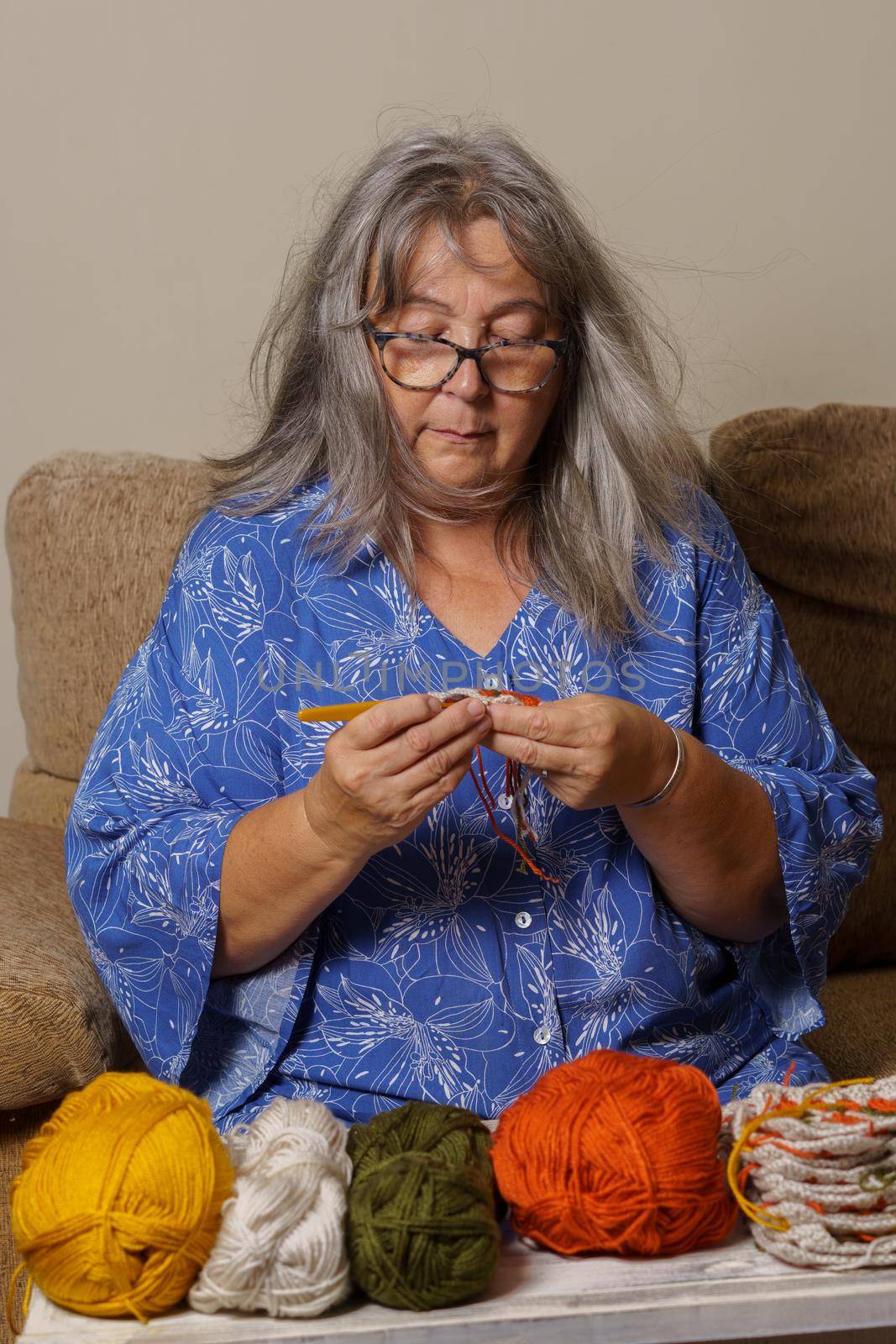 older woman with white hair and glasses crocheting on the sofa at home