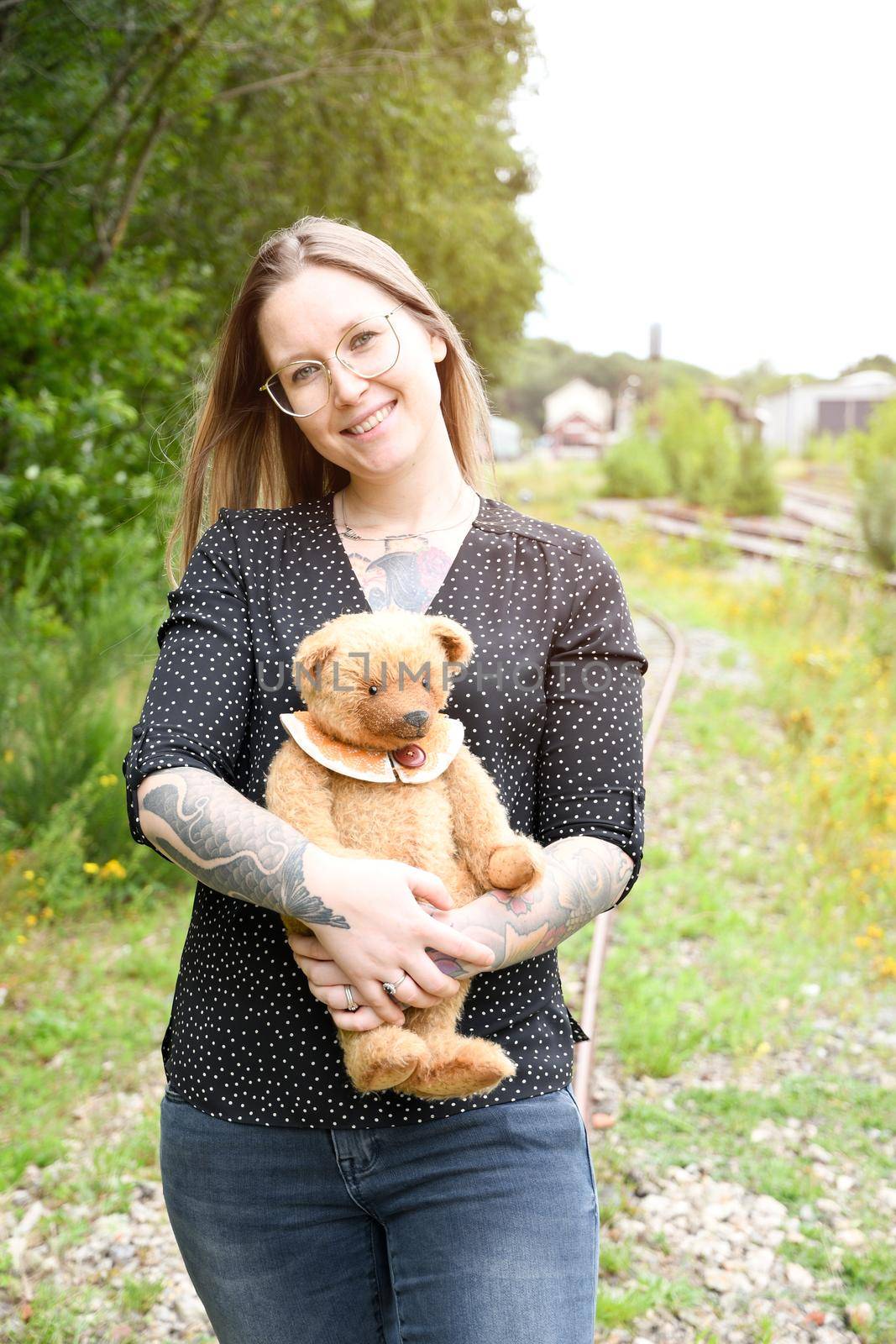 a young caucasian girl with glasses and tattoos smiles, a brown teddy bear sits in a copper pot against the background of an abandoned railway and old rails. High quality photo