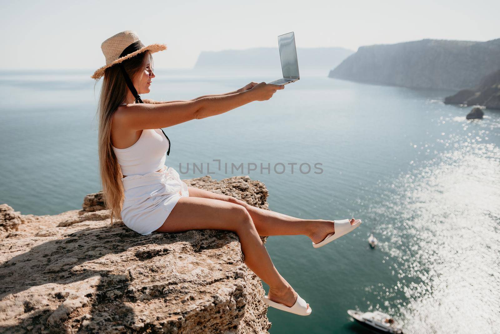 Successful business woman in yellow hat working on laptop by the sea. Pretty lady typing on computer at summer day outdoors. Freelance, travel and holidays concept.