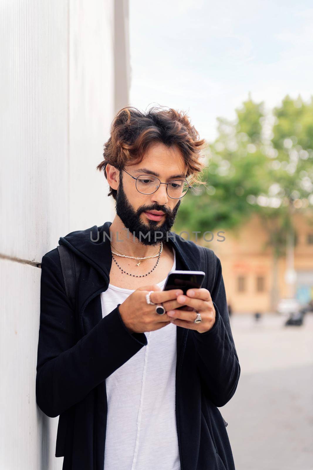 portrait of a stylish young man with beard leaning in a white wall typing on mobile phone, concept of urban lifestyle and technology of communication, copy space for text