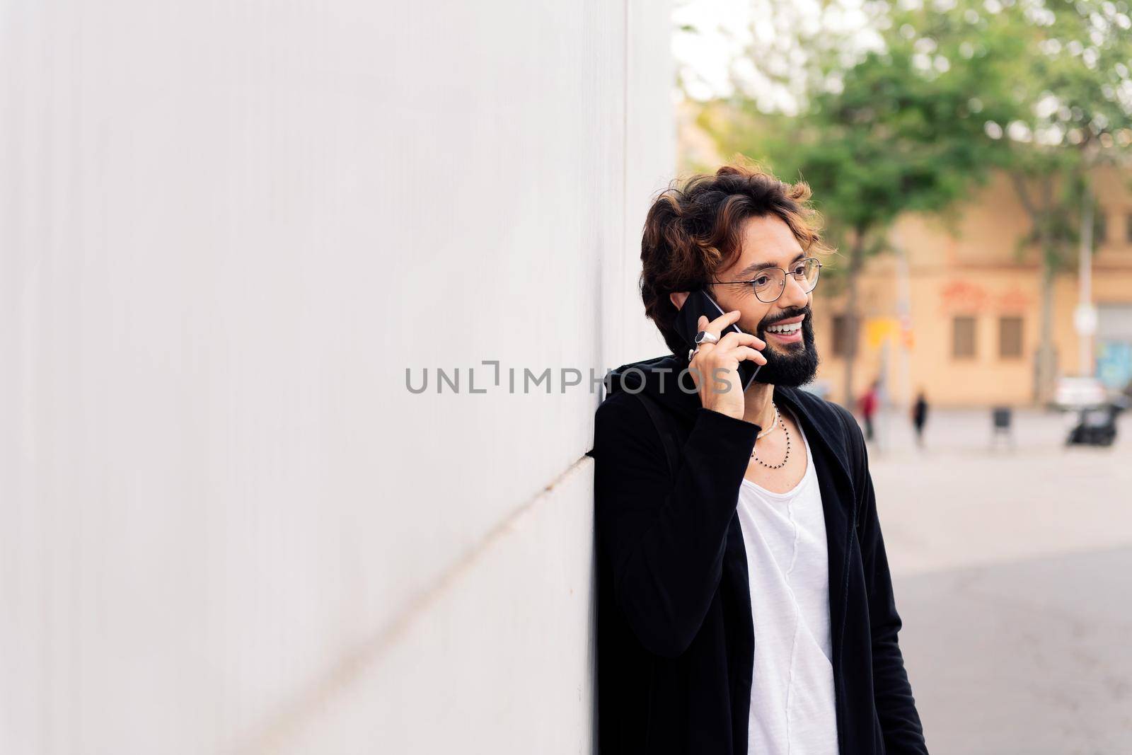 smiling young man with beard leaning in a white wall talking happy on mobile phone, concept of urban lifestyle and technology of communication, copy space for text