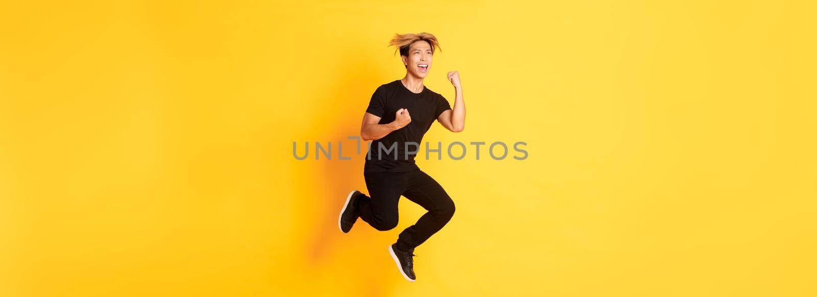 Full-length of happy attractive asian guy in black clothes jumping and celebrating victory, achieve goal, standing yellow background, triumphing.