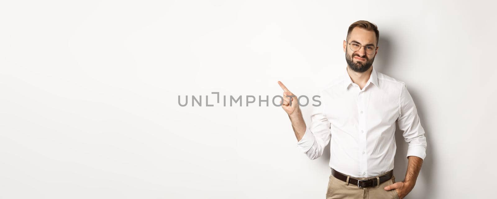 Skeptical male entrepreneur grimacing, pointing finger left with displeased face, complaining on product, white background by Benzoix