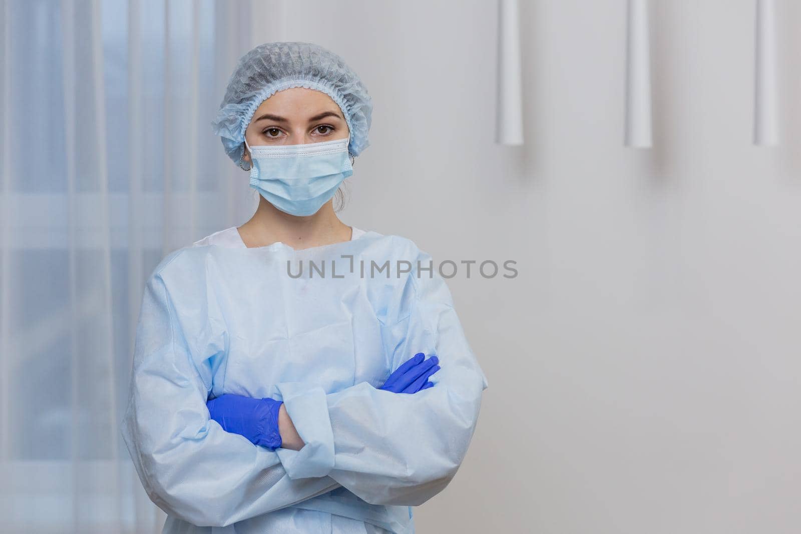Portrait of a young female doctor in a mask, surgical gown and hat, standing with arms crossed, looking at camera
