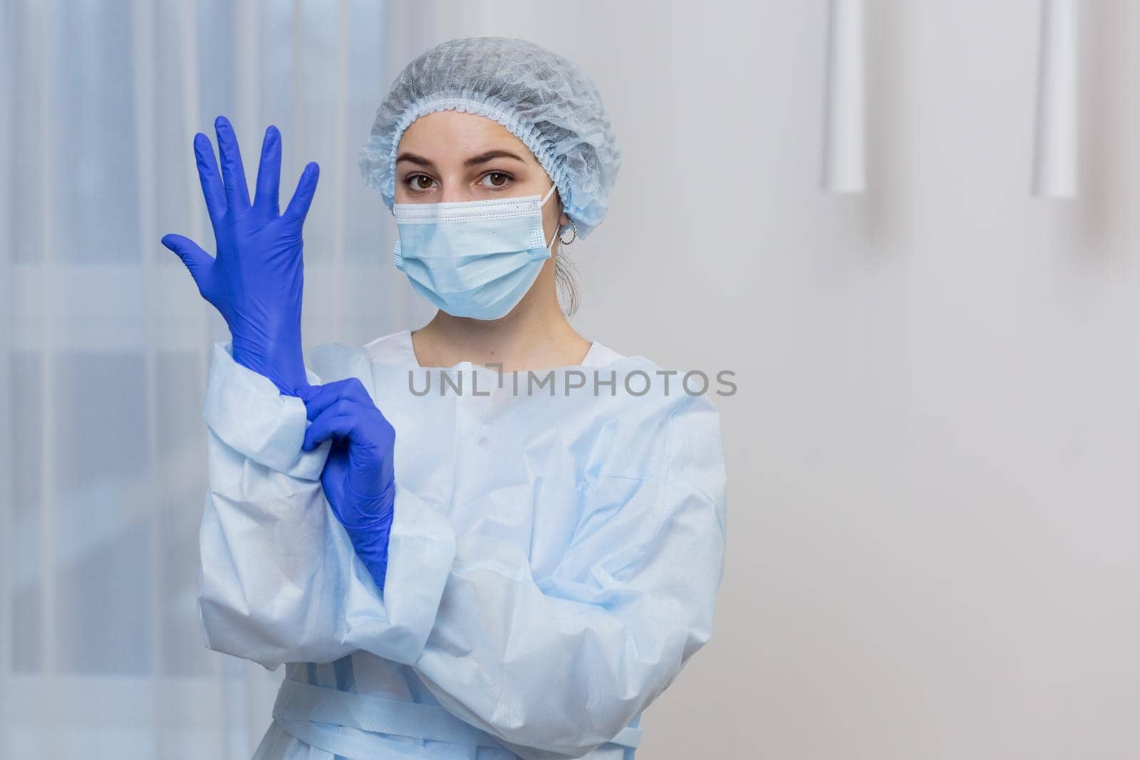 Young female doctor prepares for surgery, wears blue surgical gloves, in a white coat and mask, looks at the camera by voronaman