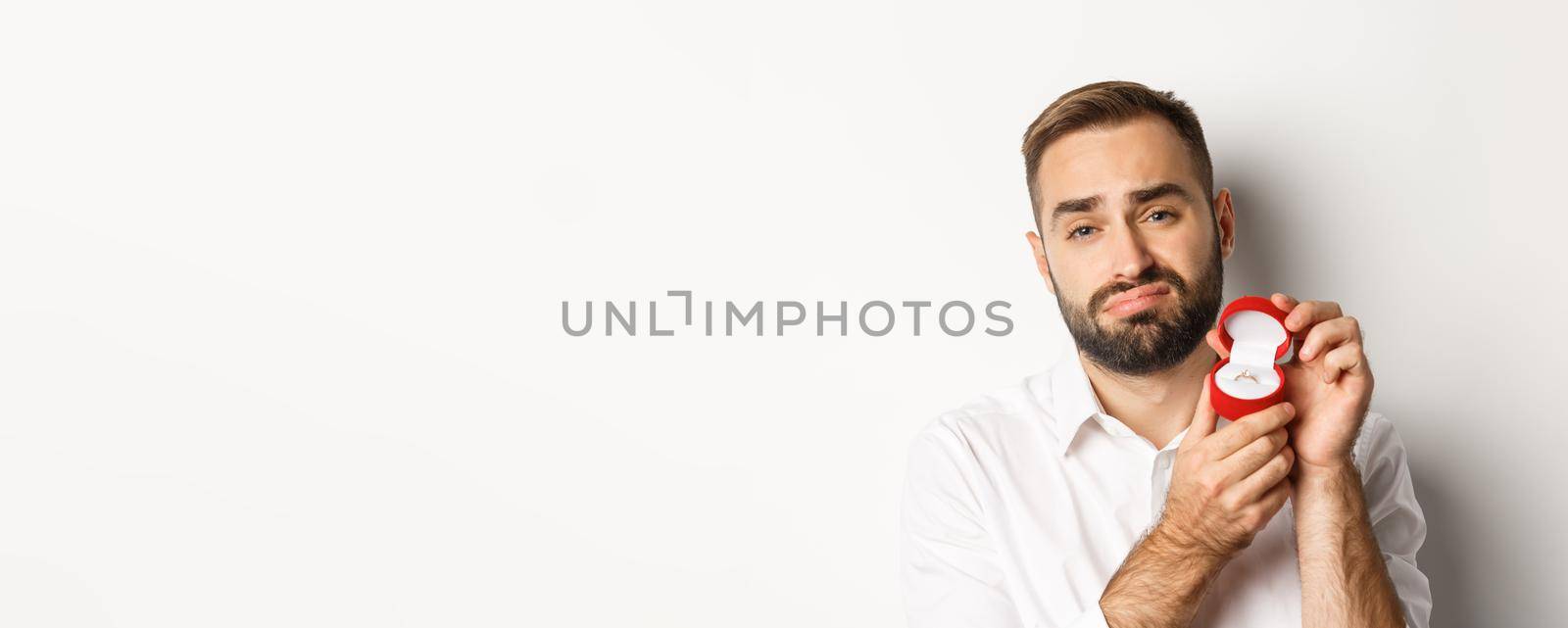 Close-up of hopeful man begging to marry him, looking sad and showing wedding ring, making a proposal, white background.