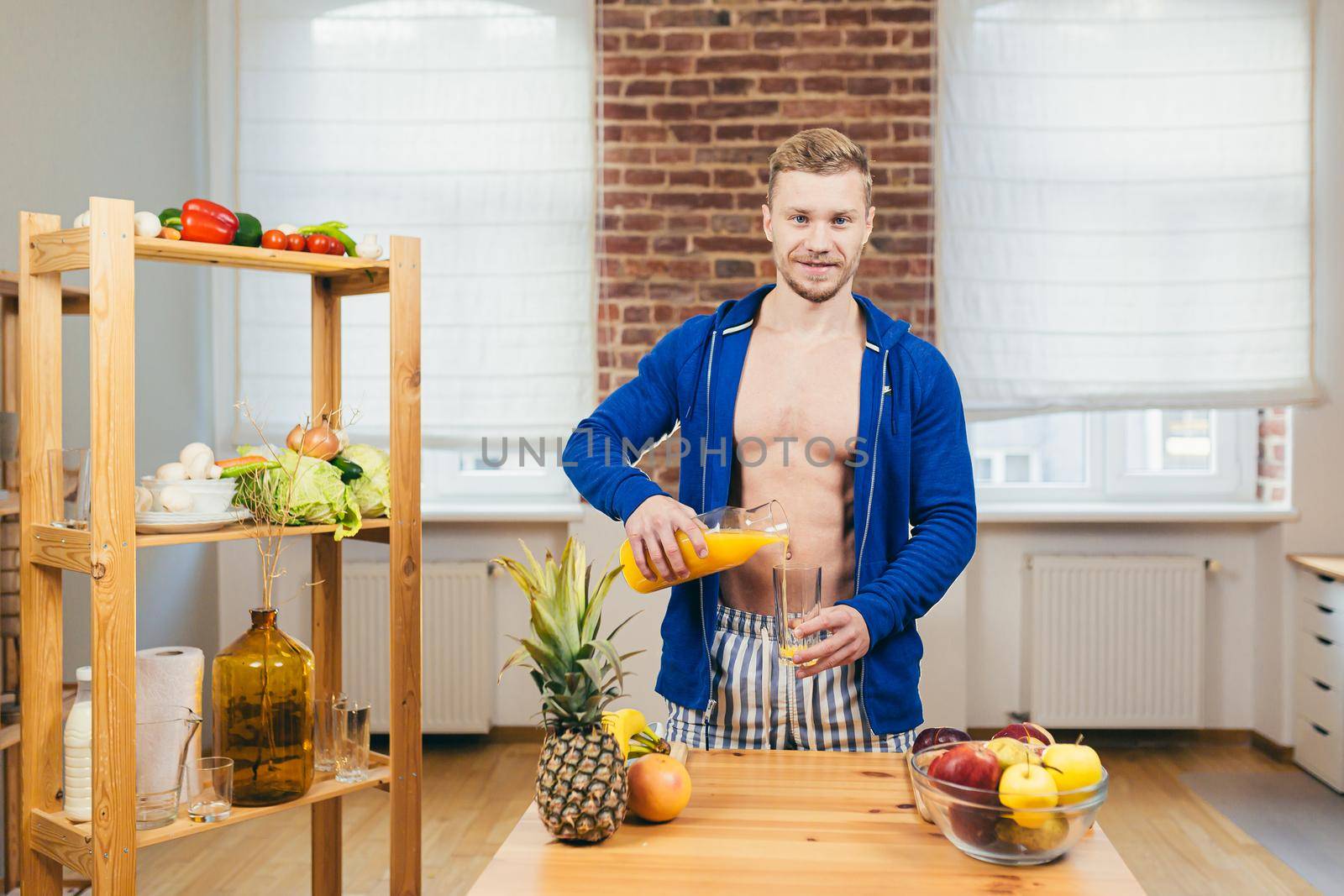 Young athletic fitness handsome man pours a glass of fresh orange juice from glass in kitchen at home, looking at camera, smiling