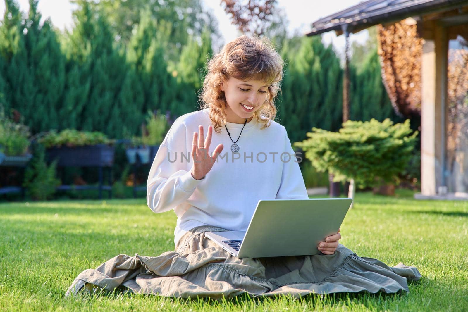 Teen girl using laptop for video communication, waving hand looking at screen, sitting on lawn green grass in backyard. Technology, chat conference video call, lifestyle, adolescence