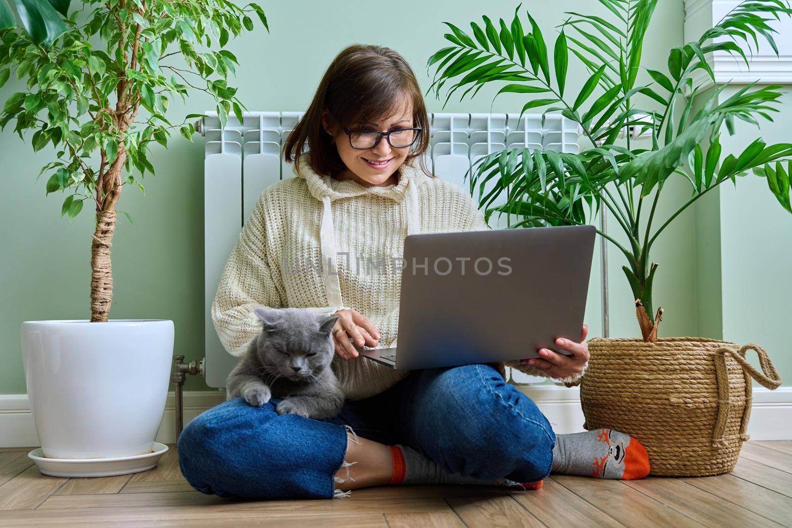 Middle-aged woman in warm clothes with cat using laptop. Female warming at home near heating radiator, with pet in her arms. Cold season winter autumn, leisure lifestyle work freelance, people animals