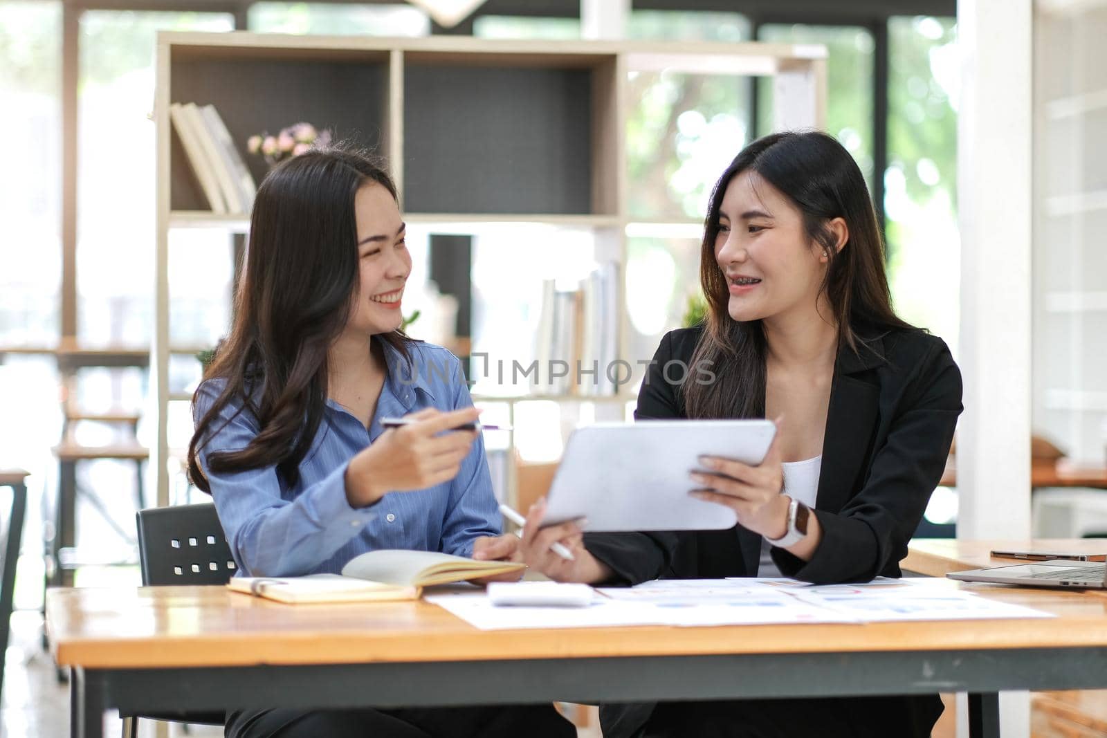 Two pretty young Asian businesswoman sitting at desk with laptop doing paperwork together discussing project financial report. Corporate business collaboration concept..