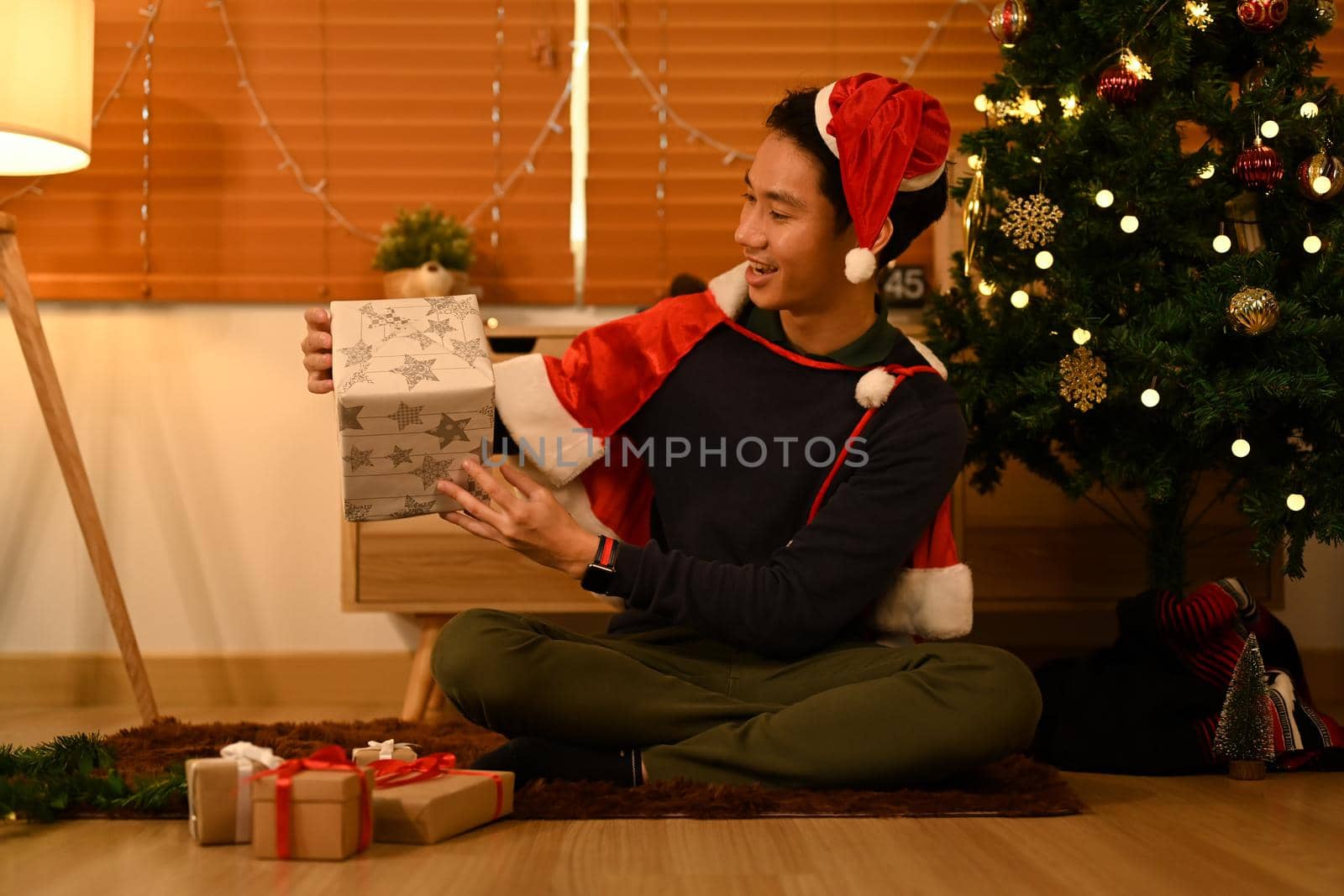 Excited man wearing Santa hat sitting near decorated Christmas tree and opening gifts. Holidays and Christmas concept.
