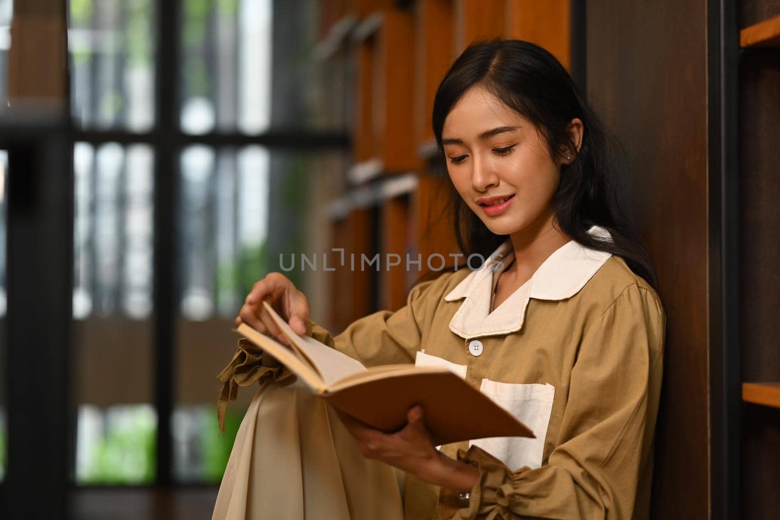 Peaceful female student reading a book between library bookshelves. Education and people concept.