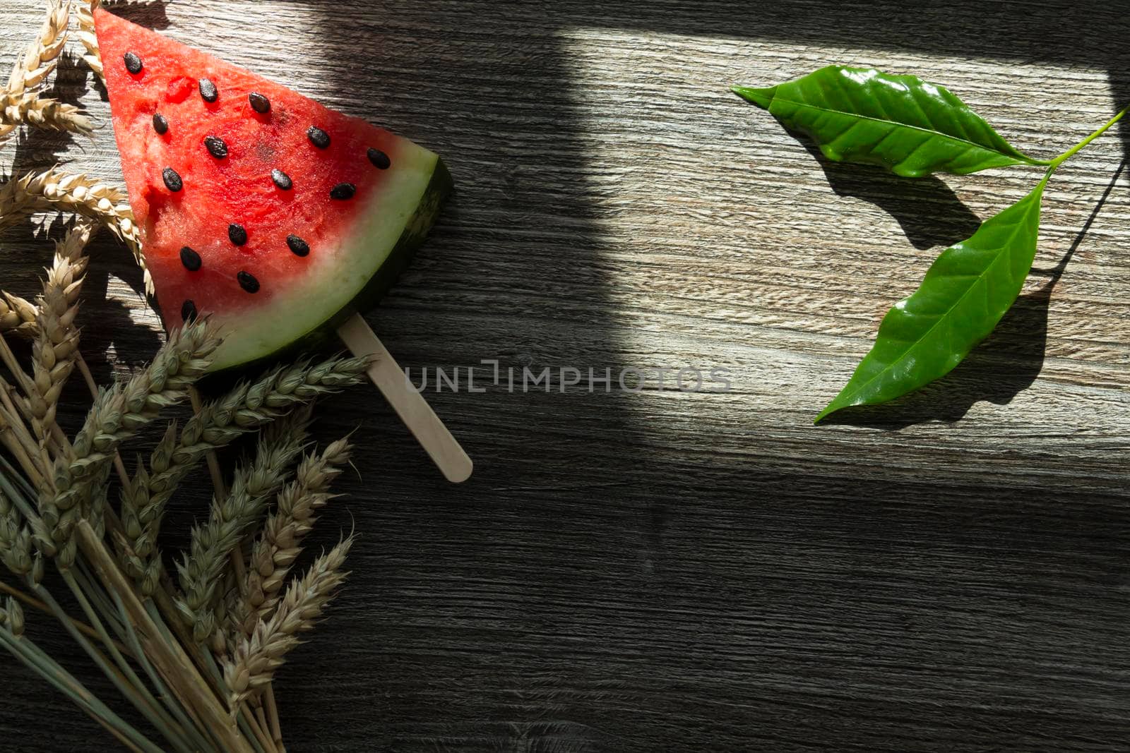 A triangular piece of sweet watermelon on an ice cream shelf, a cold drink with ice and ripe wheat ears on a wooden surface.,,