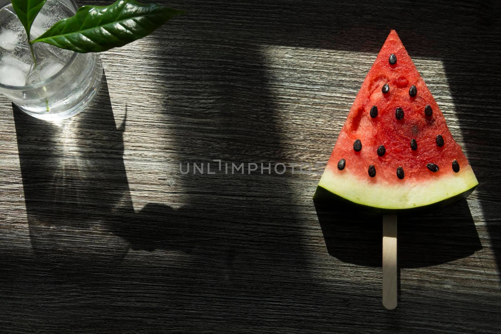 A piece of triangular watermelon on an ice cream stick and a glass of cold drink with ice.,,