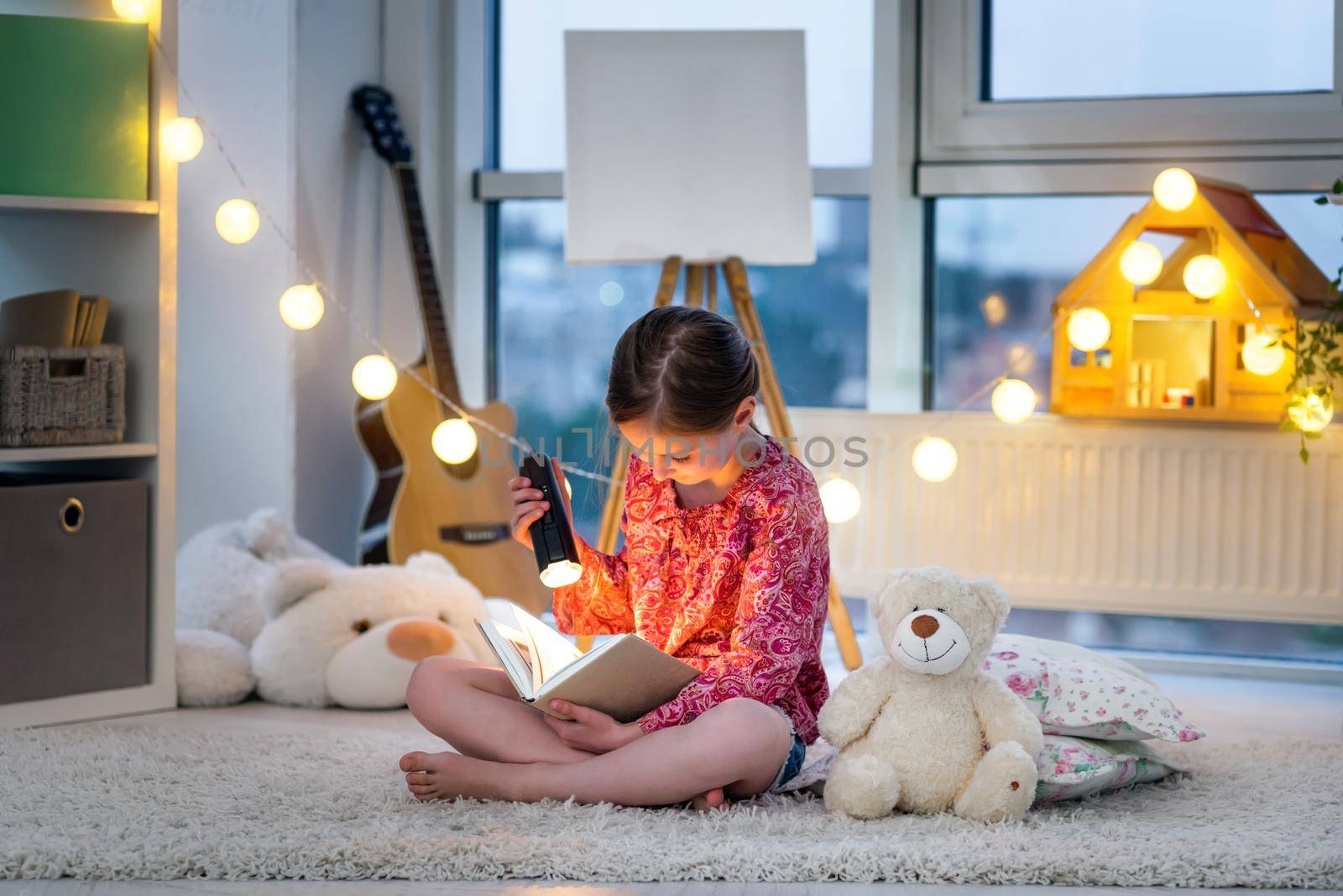 Pretty little girl with flashlight and book on floor in twilight room