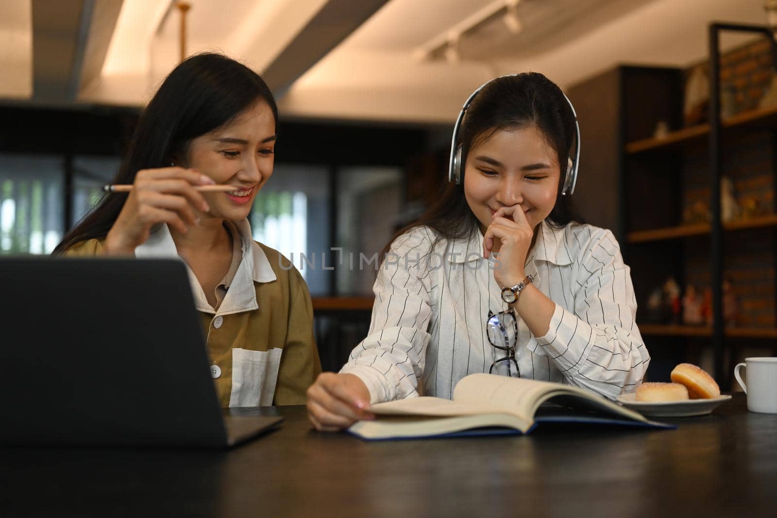 Two cheerful young students reading book while preparing for exams in college library. Education, people and technology concept.