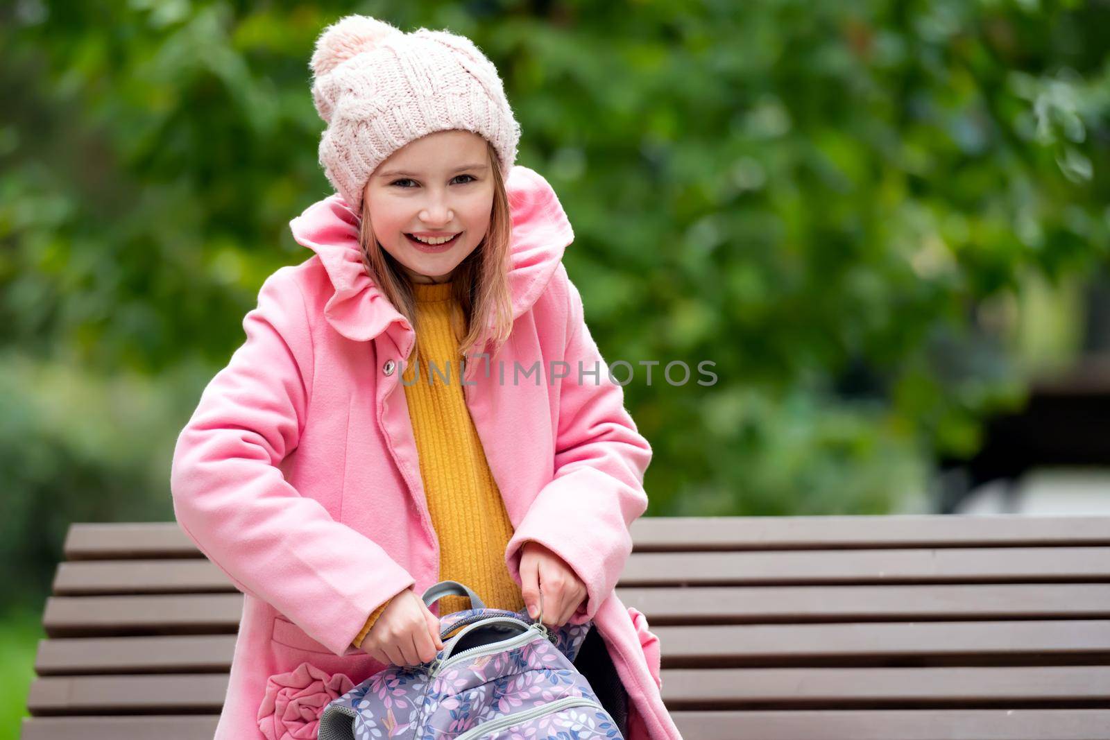 Prety school girl kid with backpack sitting on the banch at autumn park and smiling. Beautiful female child happy portrait after lessons