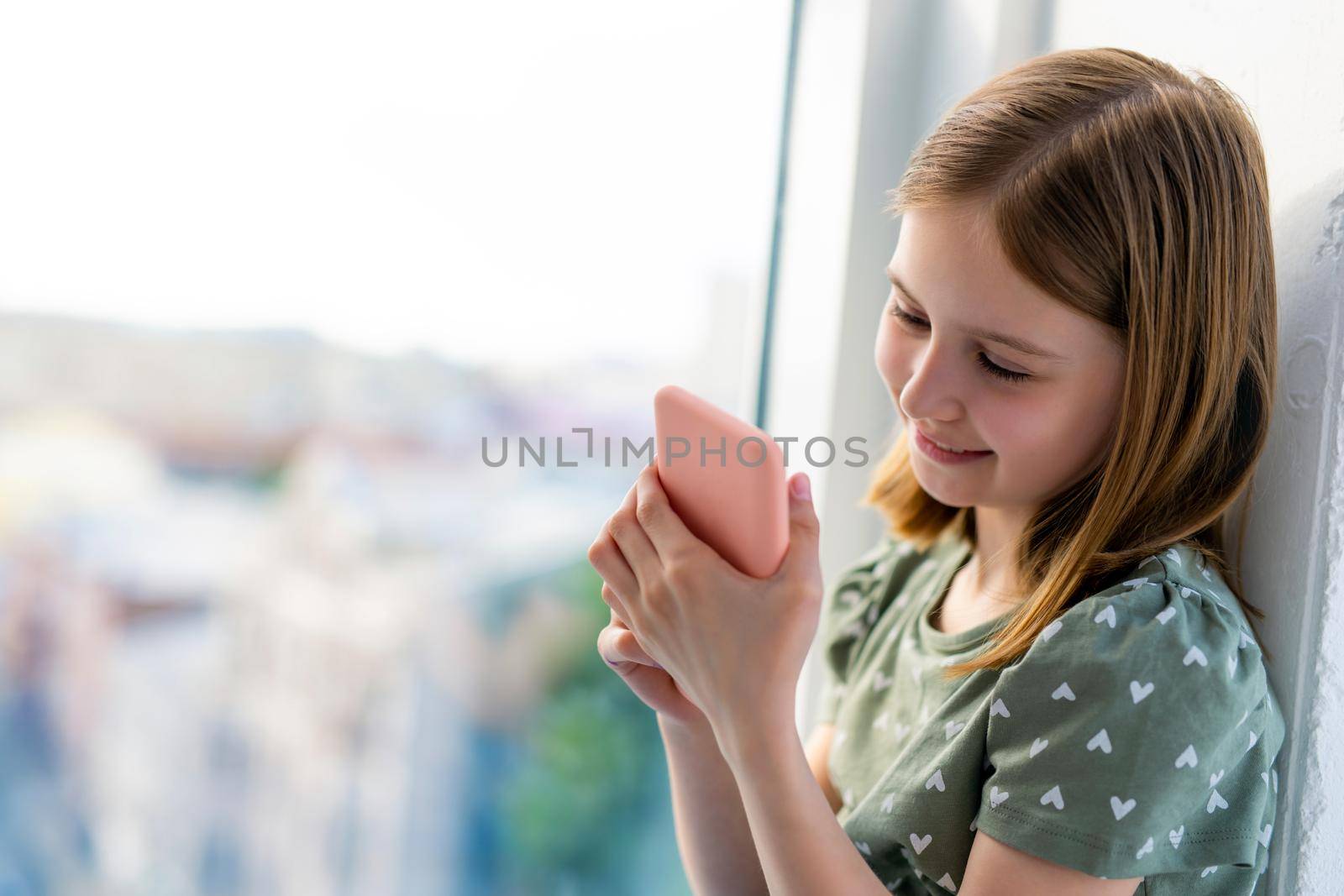 Pretty preteen girl looking at smartphone and smiling at home close to window with daylight. Beautiful kid with cell phone indoors