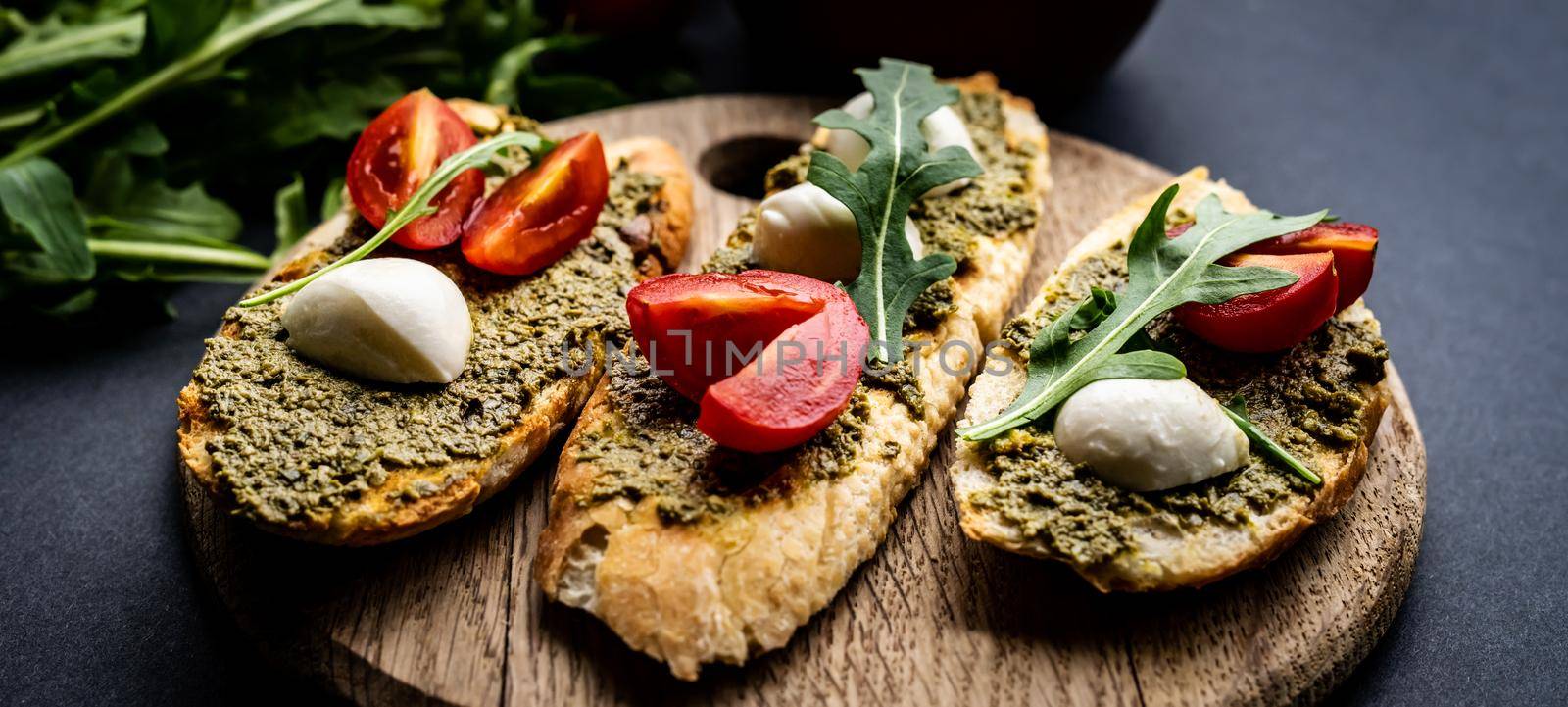 Bruschettas with pesto, tomatoes, mozzarella cheese and basil on wooden board served with guacamole, arugula and salt with herb. Italian tosted bread with vegetables and traditional sauce