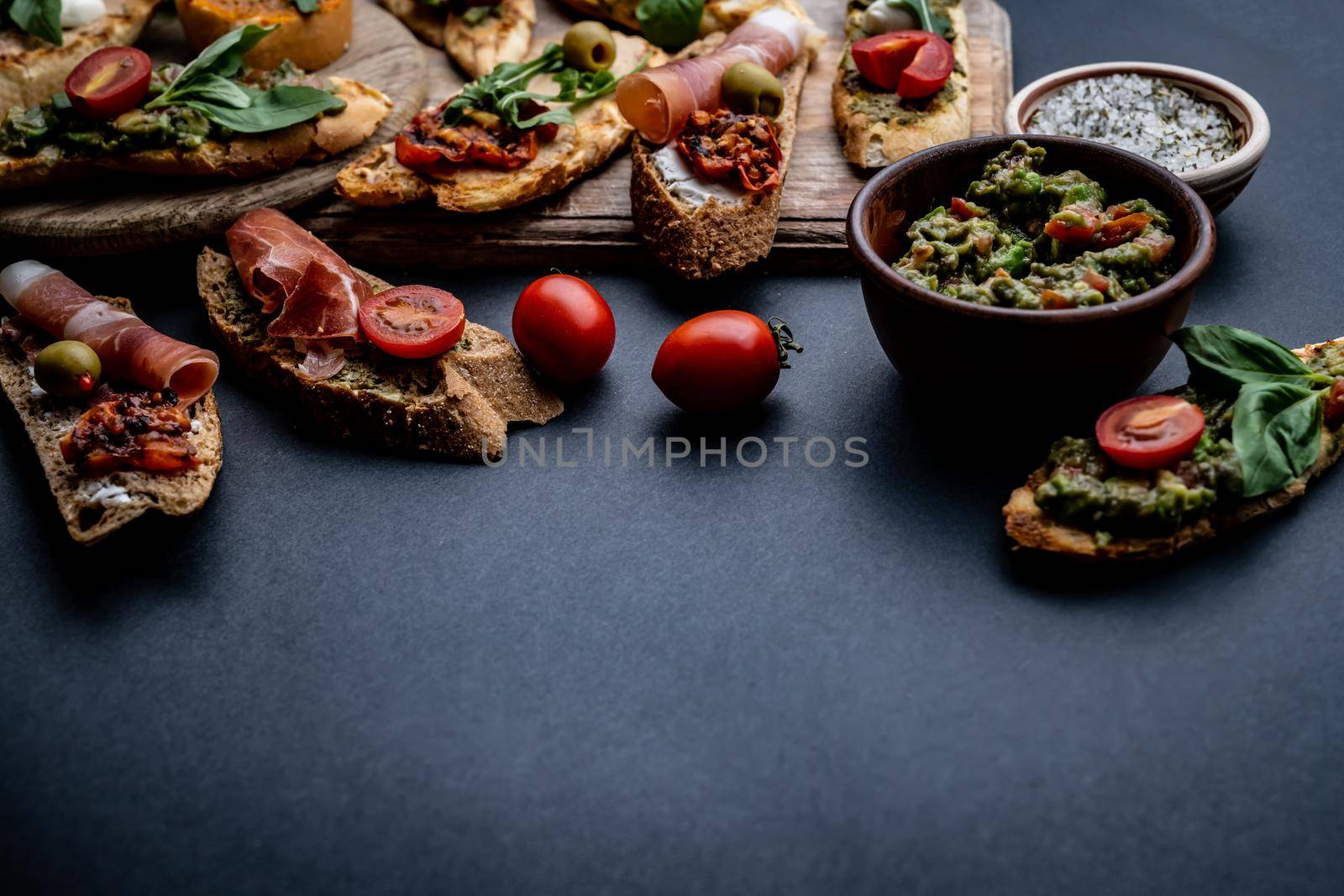 Set of bruschettas with jamon, olives, pesto, tomatoes, basil and mozzarella served on wooden board with guacamole and cherries. Traditional mediterranean antipasti food composition with copy space