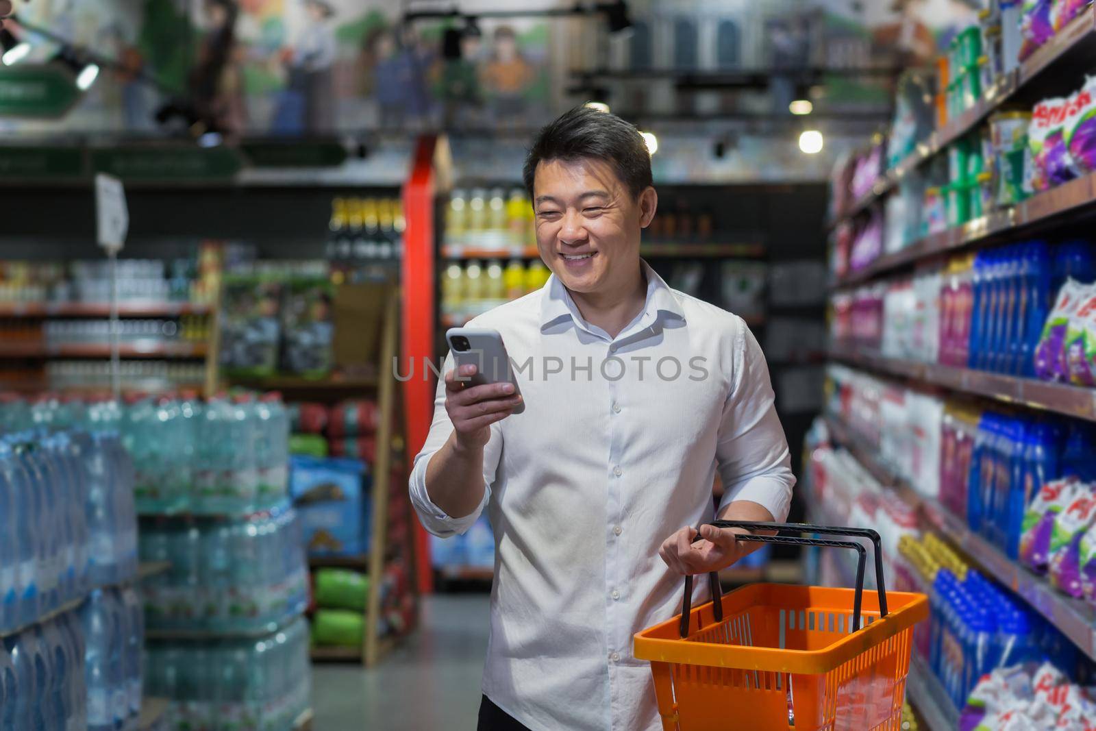 Experienced mature Asian shopper in supermarket, man smiling and reading message on smartphone, makes purchases and chooses products from various shelves