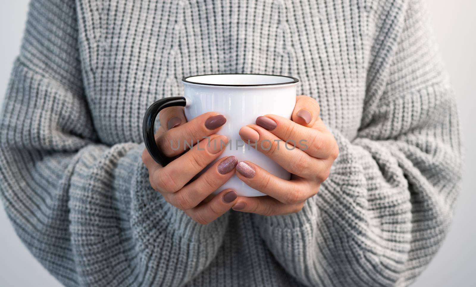 Person holding mug with hot beverage in hands. Girl in sweater with rustic cup in winter time closeup