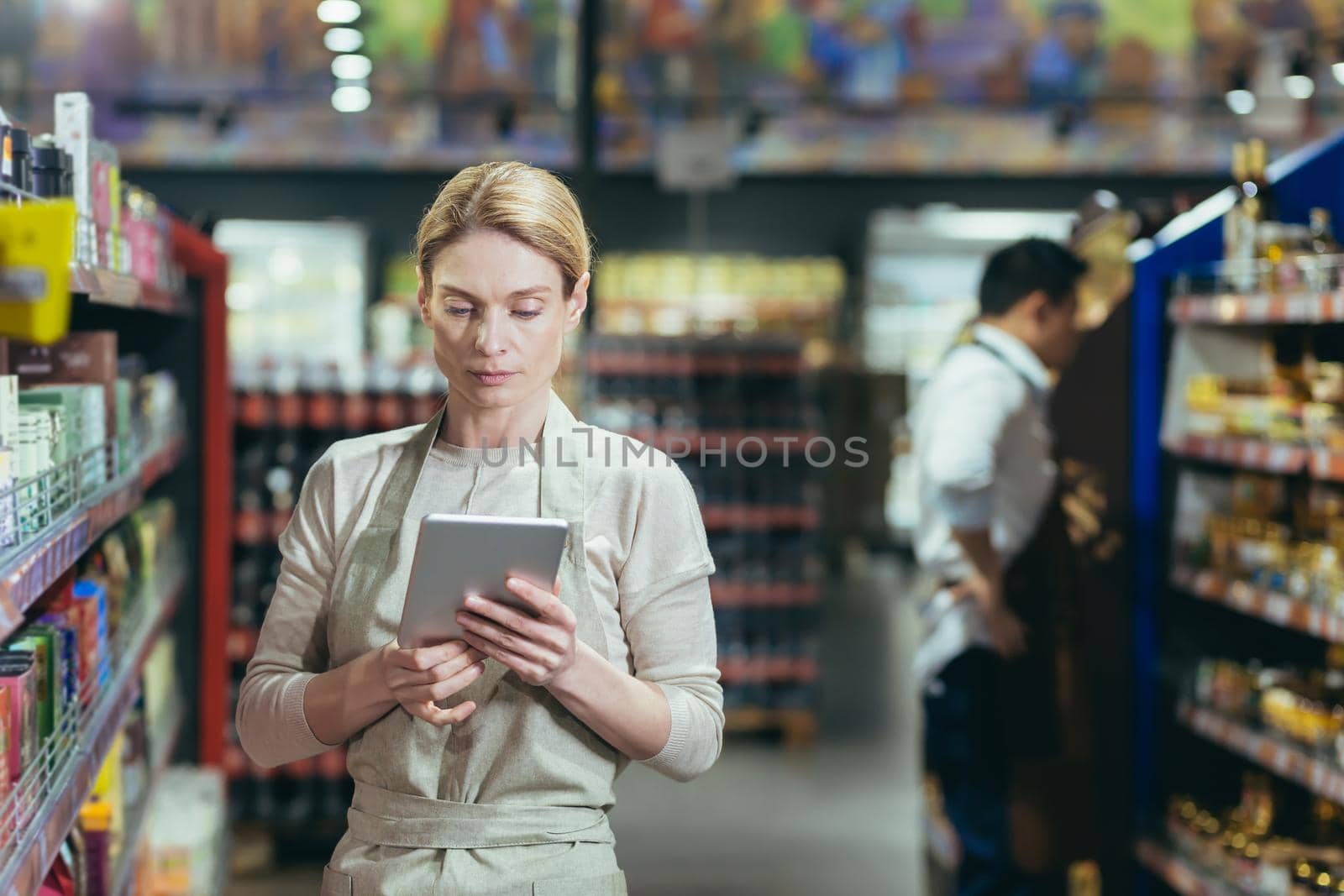 A woman seller in a supermarket uses a tablet computer to count the remaining goods, colleagues conduct an inventory in the grocery department among the shelves with goods