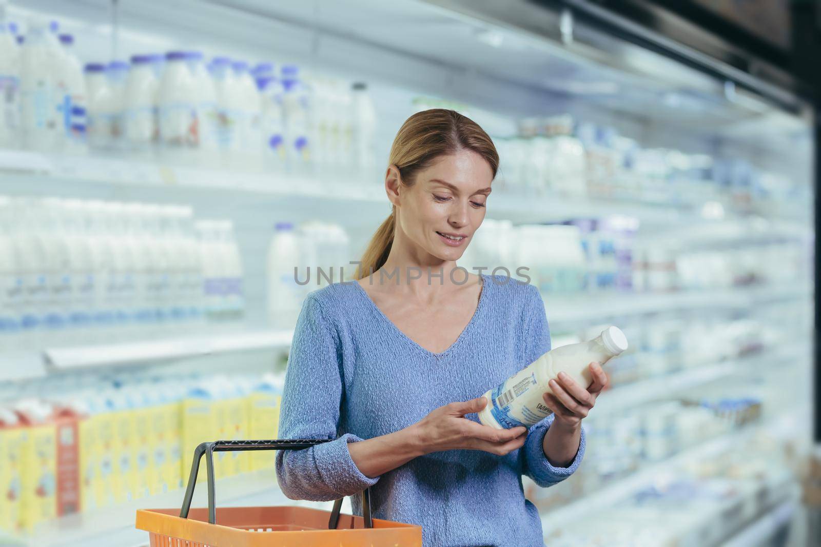 woman chooses dairy products holding a bottle of milk in her hands. female stands in a grocery store picking with a shopping basket. Studies and reads information on the label. Considers makes choices