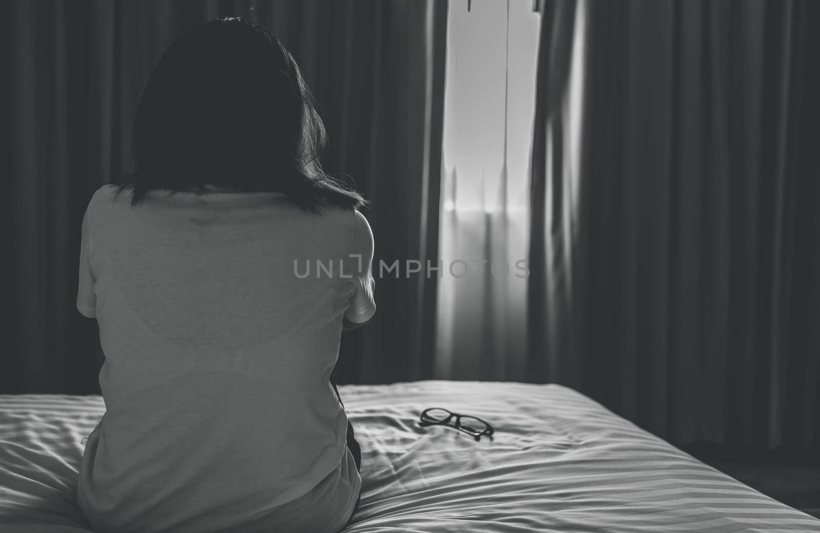 Rear view of sad and depressed woman take off glasses and sit on bed in hotel bedroom. Sad woman sit in dark bedroom. Girl with mental health problems. Unhappy life. World Mental Health Day concept.