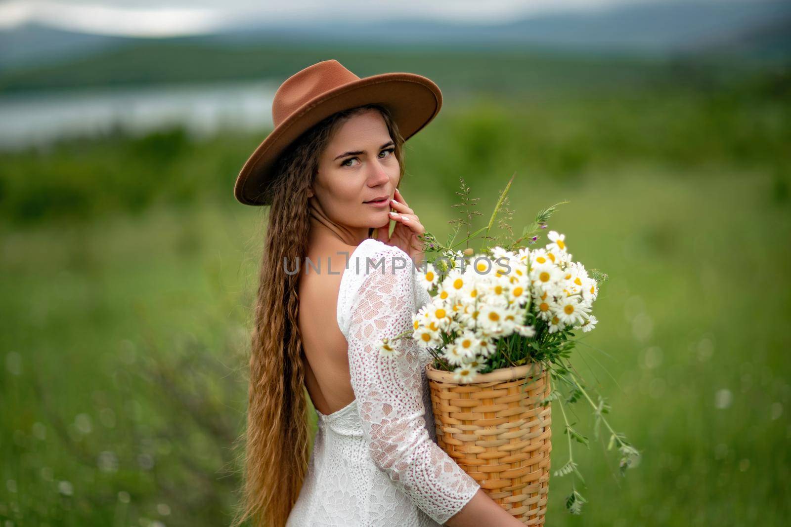 A middle-aged woman in a white dress and brown hat stands on a green field and holds a basket in her hands with a large bouquet of daisies. In the background there are mountains and a lake