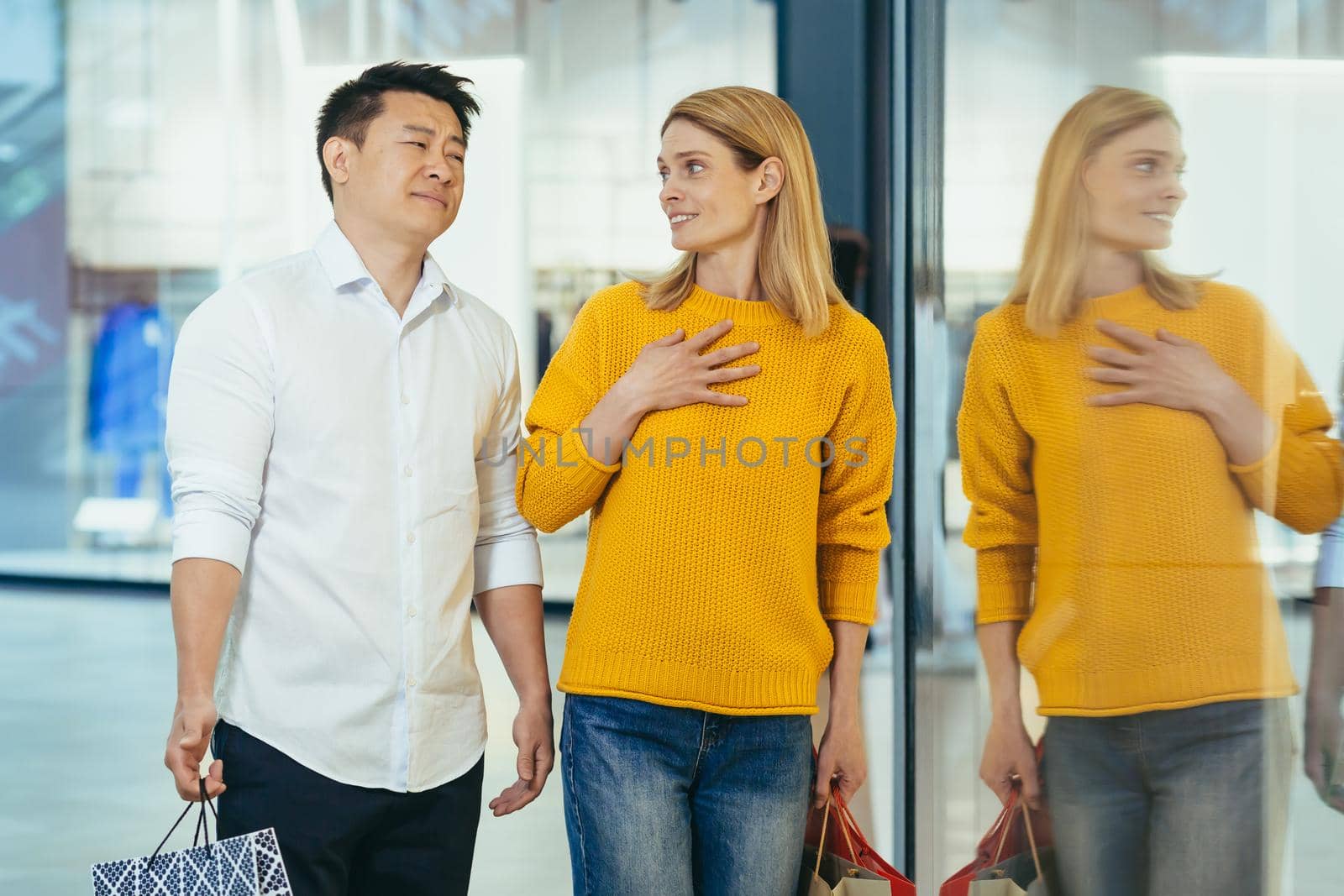 multiracial couple asian man and caucasian woman walking together in mall after shopping with colored bags in hands. walking past shop windows with clothes cheerful family hugging smile. Asia by voronaman