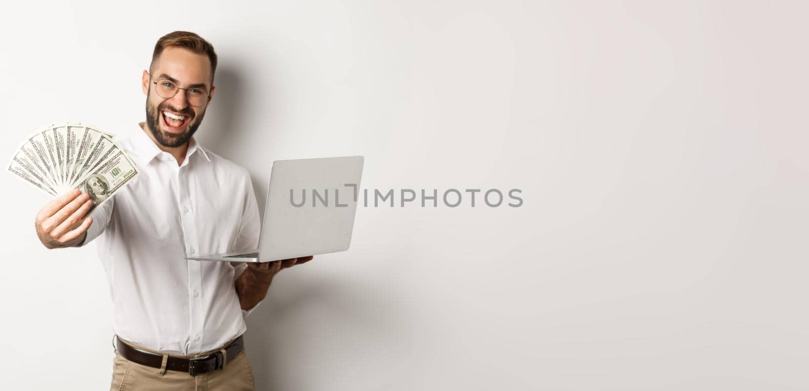 Business and e-commerce. Happy successful businessman bragging with money, working on laptop online, standing over white background by Benzoix