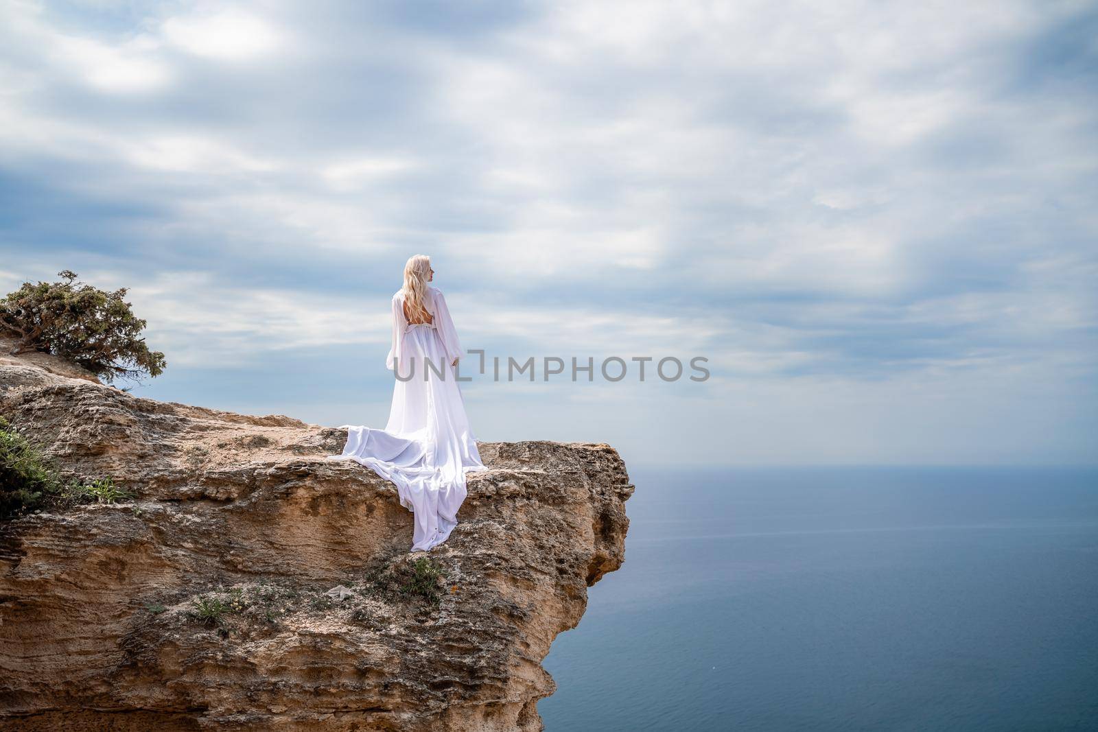 Blonde with long hair on a sunny seashore in a white flowing dress, rear view, silk fabric waving in the wind. Against the backdrop of the blue sky and mountains on the seashore