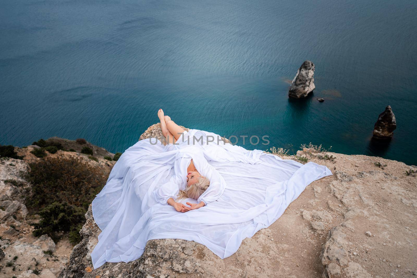 The blonde lies on a rock above the sea. With long hair on a sunny seashore in a white dress, rear view, silk fabric flutters in the wind. Against the backdrop of blue skies and mountains