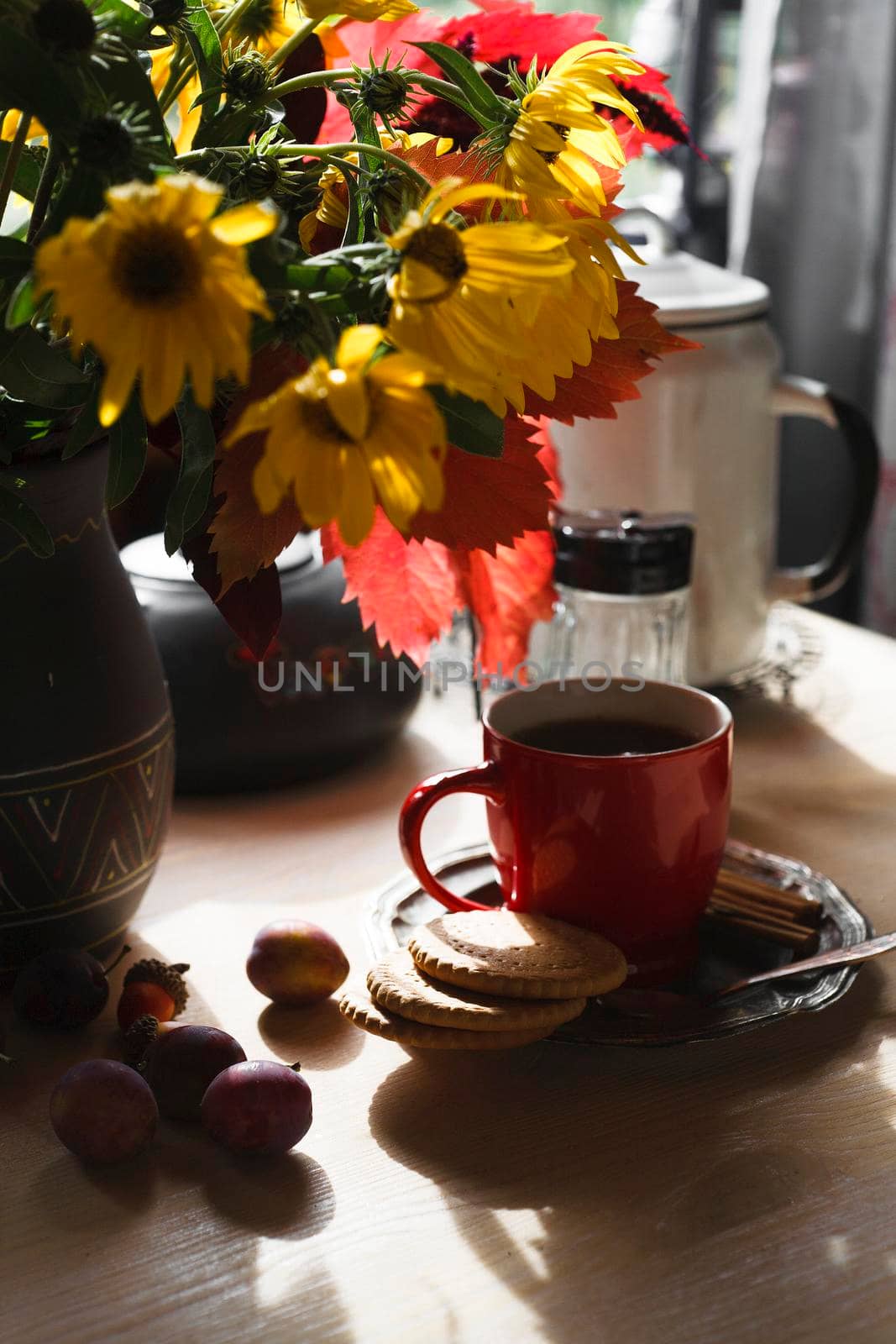 Red cup of tea on metal plate with a bouquet of yellow autumn flowers and colored leaves and plums on white table, early autumn morning concept, selective focus.