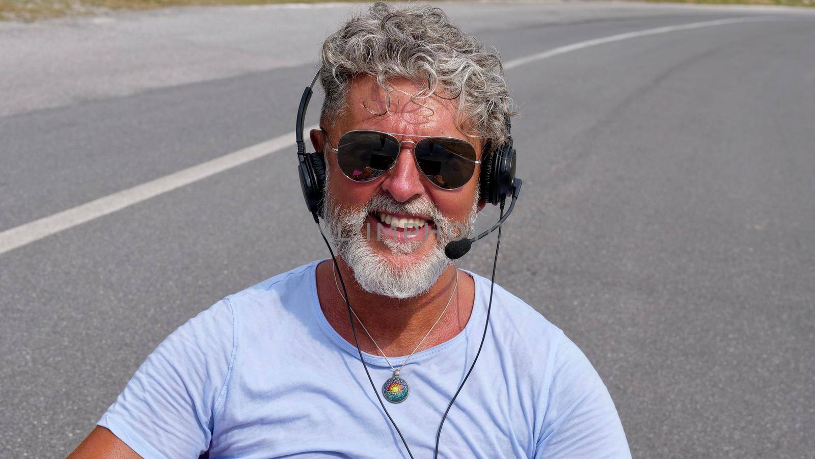 Gray-haired elderly man freelancer with a beard in sunglasses, working in the headphones on road. A crazy and extraordinary old senior in the summer