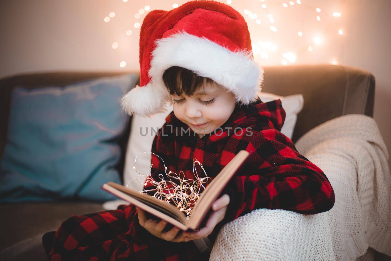 A boy in a Santa hat is reading a book on a lifestyle sofa. Reading books. New Year's mood. Garland on the wall