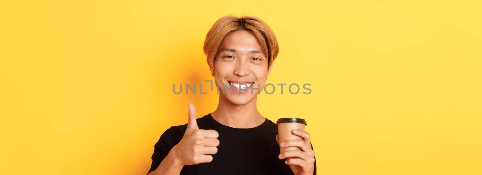 Close-up of satisfied asian handsome guy recommend cafe, holding cup of coffee and showing thumbs-up in approval, smiling pleased over yellow background.