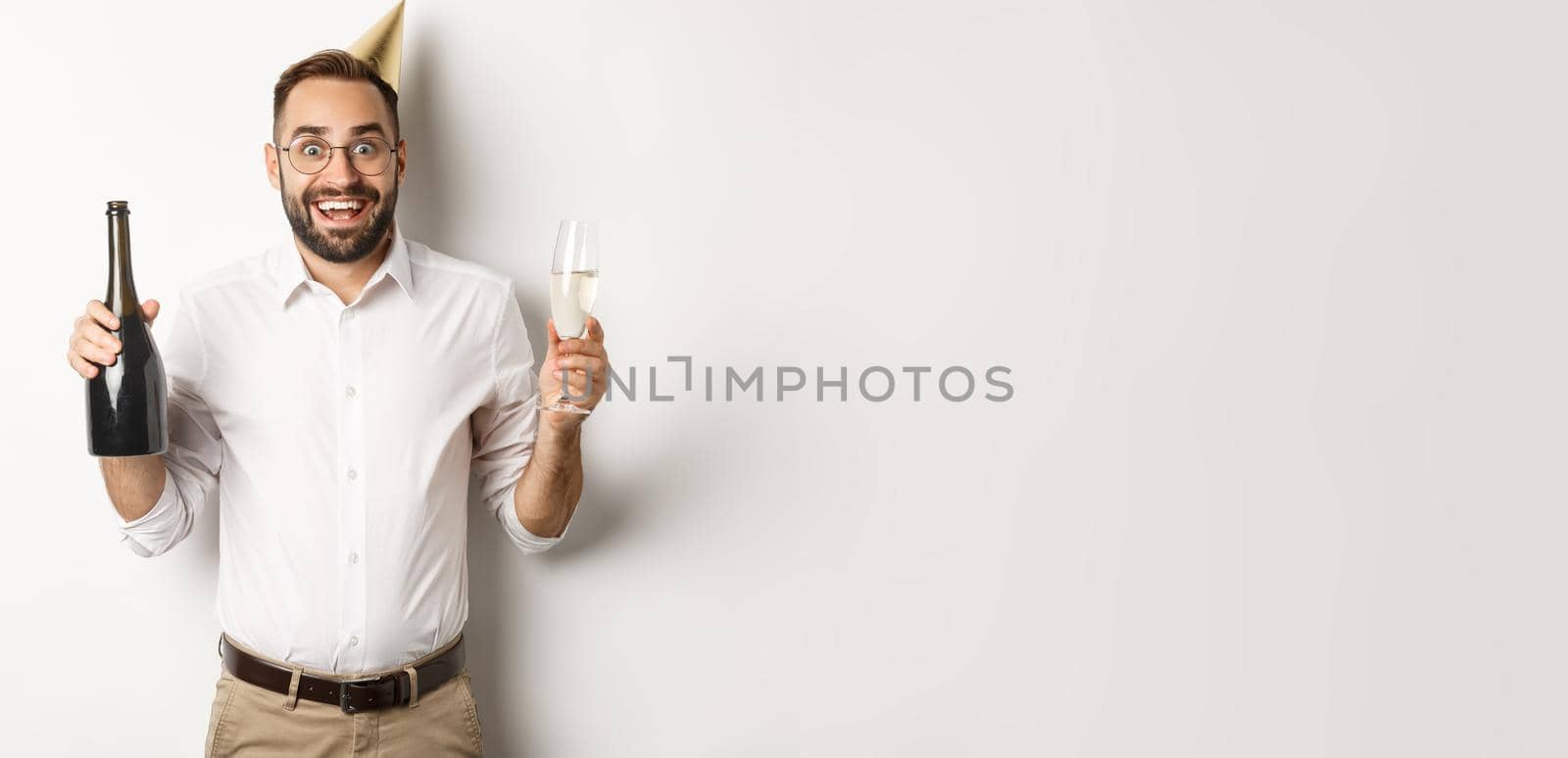 Celebration and holidays. Excited man enjoying birthday party, wearing b-day hat and drinking champagne, standing over white background.