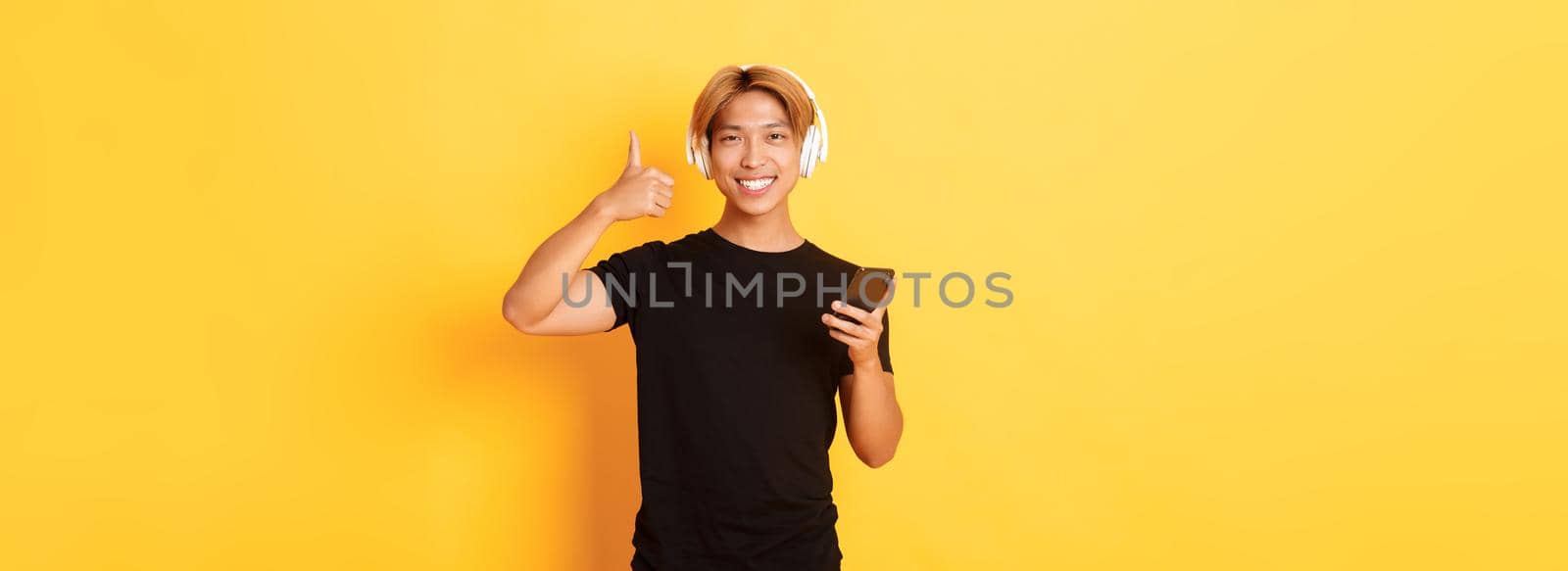 Happy satisfied asian guy likes music or podcast, showing thumbs-up in approval, holding mobile phone, standing yellow background.