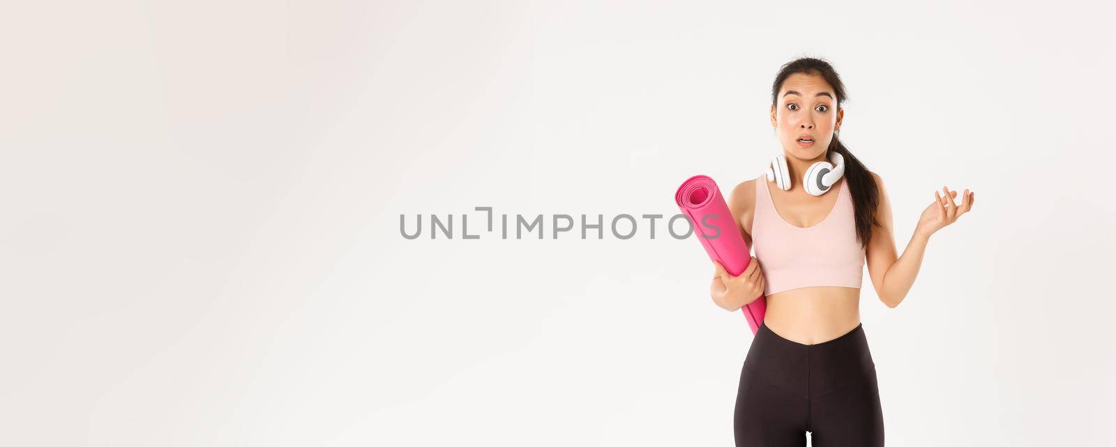 Sport, wellbeing and active lifestyle concept. Confused and shocked asian fitness girl, cant understand why gym closed, holding rubber mat and looking surprised at camera, white background by Benzoix