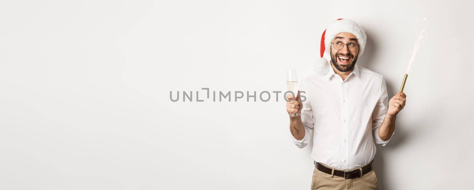 Winter holidays and celebration. Happy businessman enjoying New Year party, wearing Santa hat and drinking champagne, smiling amused, white background.